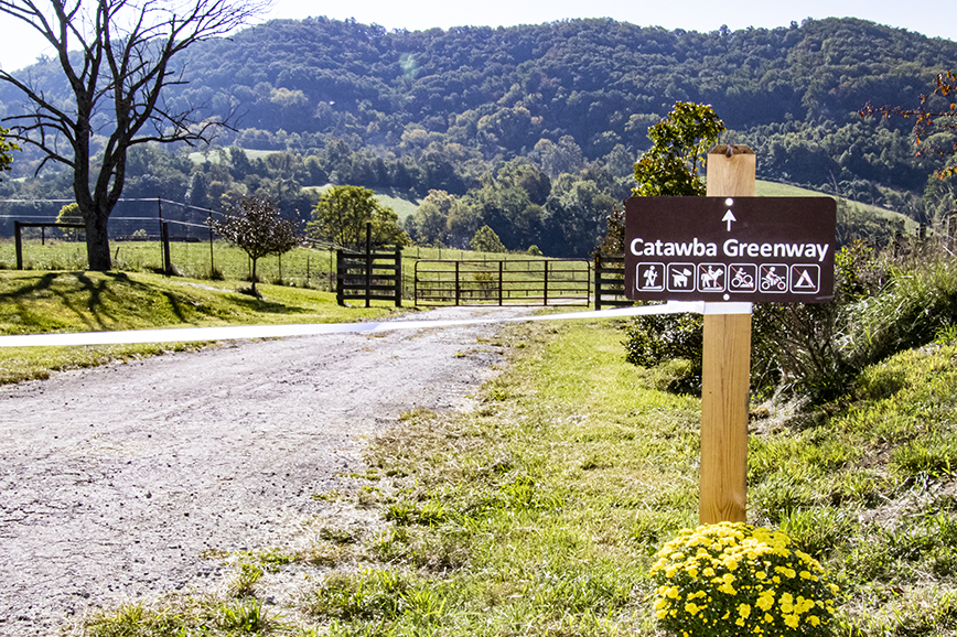 A brown Catawba Greenway sign next to a gravel road that leads to mountains in the distance.