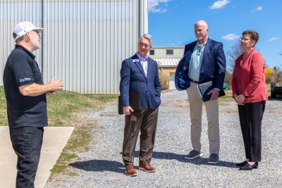 Four people stand together on a gravel driveway talking with a blue spring sky overhead.