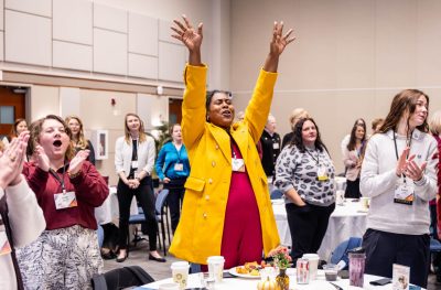 A woman in a bright yellow suit raises her hands in the air and cheers while others cheer around her.