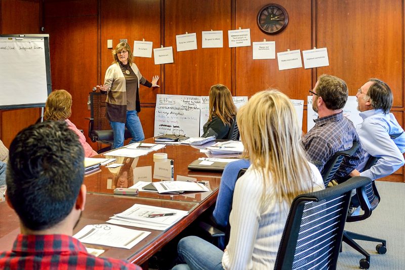 A woman stands at the front of the room next to large white papers with writing taped to the wall. Students sit around a board table in front of her.