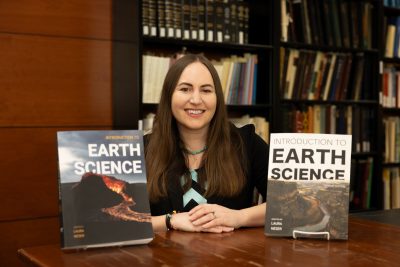 A woman with long brown hair is seated at a wooden table in a library, smiling at the camera. She is wearing a black blazer and turquoise necklace. In front of her are two copies of her textbook titled "Introduction to Earth Science", one with a volcanic landscape on the cover and the other with a river canyon scene. Bookshelves filled with books are visible in the background.