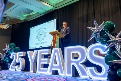 Ken Nicely speaking on stage in front of a large lighted sign that says "25 years"