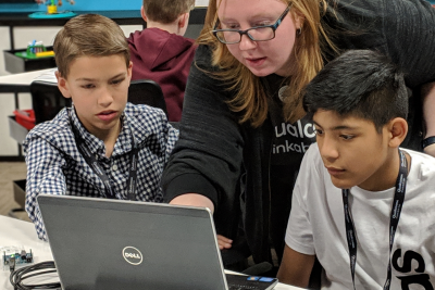 A teacher leans over the shoulders of two boys to point at something on the screen of a laptop in front of them.