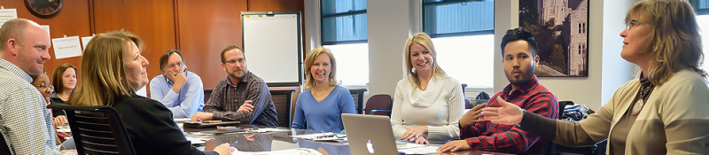 A teacher and adult students sit at a long conference table.