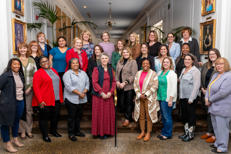 Outside group photo of about 25 women with dark woods in the background.